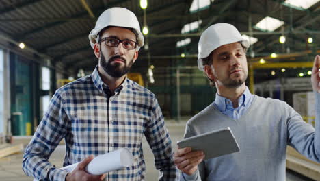 Two-Caucasian-men-wearing-helmets-walking,-talking-and-looking-at-the-tablet-screen-in-a-factory