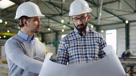 Two-Caucasian-men-wearing-helmets-talking-and-looking-at-a-blueprint-in-a-factory