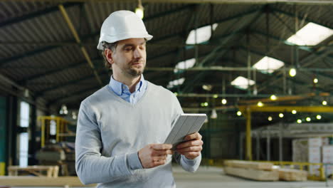 Caucasian-male-worker-wearing-helmet-and-walking-while-holding-a-tablet-and-looking-around-in-a-factory
