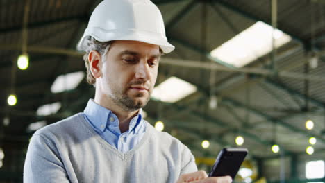 Close-up-view-of-caucasian-male-worker-wearing-a-helmet-typing-on-smartphone-in-a-factory