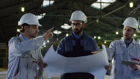 Three-engineers-wearing-helmets-holding-blueprint-and-tablet-while-talking-and-walking-in-a-factory