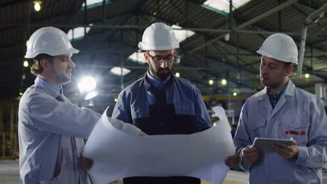 Three-engineers-wearing-helmets-holding-blueprint-and-tablet-while-talking-in-a-factory