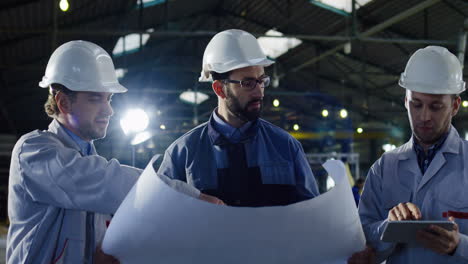Three-engineers-wearing-helmets-holding-blueprint-and-tablet-while-talking-in-a-factory