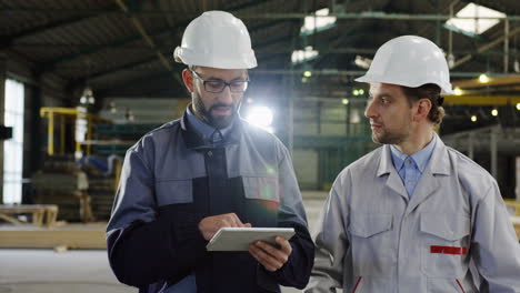 Two-engineers-wearing-helmets-holding-tablet-while-talking-and-walking-in-a-factory