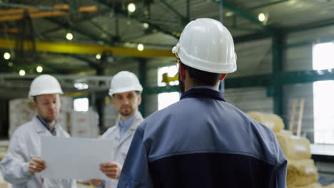 Rear-view-of-the-male-factory-worker-wearing-a-helmet-and-approaching-two-coworkers