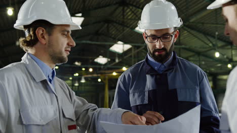 Close-up-view-of-three-engineers-wearing-helmets-and-holding-blueprint-while-talking-in-a-factory