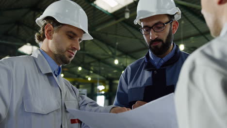 Close-up-view-of-three-engineers-wearing-helmets-and-holding-blueprint-while-talking-in-a-factory