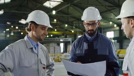 Three-engineers-wearing-helmets-and-holding-blueprint-while-talking-in-a-factory