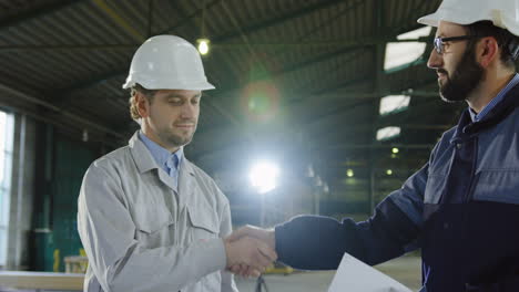 Two-engineers-wearing-helmets-and-holding-blueprint-while-talking-and-shaking-hands-in-a-factory