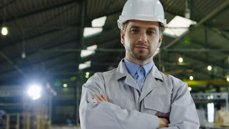 Caucasian-engineer-wearing-a-helmet-turning-his-head-to-the-camera,-smiling-and-crossing-arms-in-a-factory
