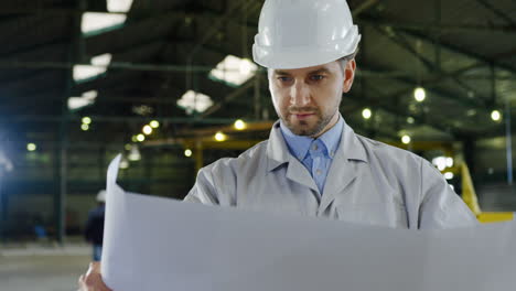 Caucasian-engineer-wearing-a-helmet-holding-blueprint-and-looking-around-in-a-factory