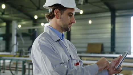 Side-view-of-engineer-wearing-a-helmet-and-typing-on-the-tablet-in-a-big-factory