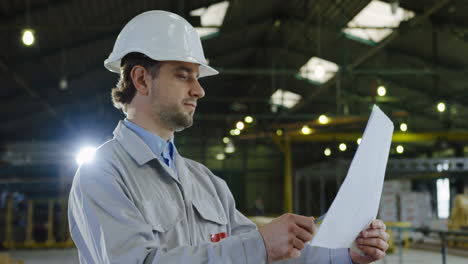 Caucasian-engineer-wearing-helmet-and-looking-around-and-at-blueprint-in-a-big-factory