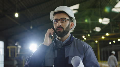 Caucasian-factory-worker-wearing-a-helmet-and-glasses,-holding-a-blueprint-and-talking-on-the-phone-in-a-big-factory