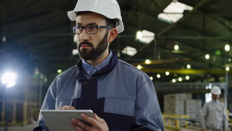 Caucasian-factory-worker-wearing-a-helmet-and-glasses,-typing-on-tablet-in-a-big-factory