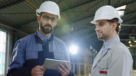 Two-workers-wearing-helmets-talking-while-looking-at-the-tablet-in-a-big-factory,-then-they-look-at-the-camera