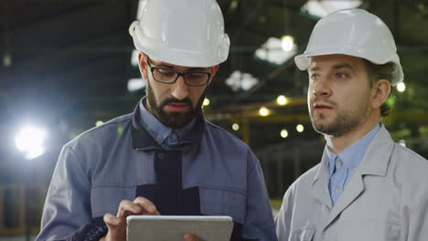Two-workers-wearing-helmets-talking-while-looking-at-the-tablet-in-a-big-factory