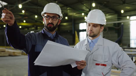Two-workers-wearing-helmets-talking-while-looking-at-blueprint-in-a-big-factory