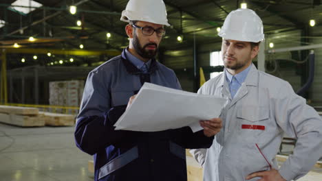 Two-workers-wearing-helmets-talking-while-looking-at-blueprint-in-a-big-factory