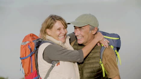 Front-view-of-a-happy-senior-hikers-with-backpacks-hugging-and-kissing,-then-looking-at-the-camera