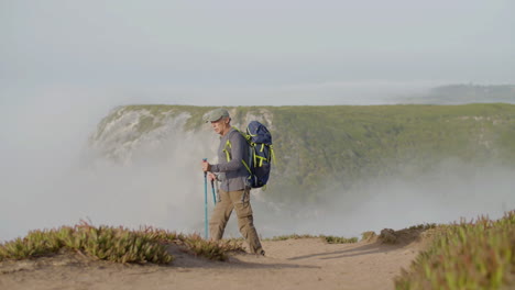 Long-shot-of-focused-man-with-backpack-climbing-mountain-with-trekking-poles-alone-on-a-foggy-day