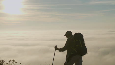 Senior-man-climbing-mountain-with-trekking-sticks-at-sunset