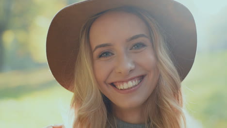 Close-up-view-of-young-blonde-woman-wearing-a-hat-and-smiling--to-the-camera-in-a-park-in-the-morning