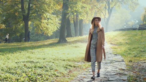 Full-lenght-view-of-caucasian-woman-with-blonde-hair-wearing-a-coat-and-hat-walking-in-the-park-in-the-morning-in-autumn