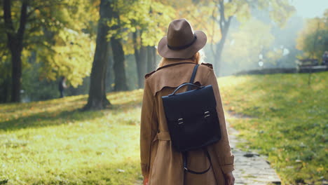 Rear-view-of-Caucasian-woman-in-coat,-hat-and-with-a-backpack-walking-in-the-park-and-looking-at-the-sides-in-autumn