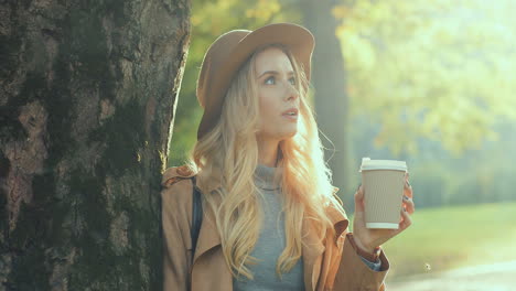 Close-up-view-of-blonde-woman-wearing-hat,-leaning-on-a-tree,-drinking-a-coffee-to-go,-and-looking-around-with-a-smile-on-her-face-in-the-park-in-autumn