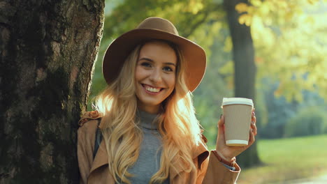 Close-up-view-of-blonde-woman-wearing-hat,-leaning-on-a-tree,-drinking-a-coffee-to-go,-and-looking-at-the-camera-with-a-smile-on-her-face-in-the-park-in-autumn