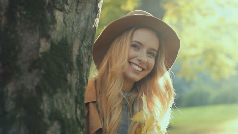 Caucasian-young-blonde-woman-wearing-hat,-holding-a-yellow-leaf-and-looking-at-camera-behind-a-tree-in-the-park-in-autumn