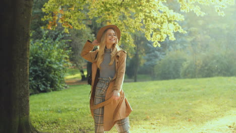 Cheerful-blonde-young-woman-wearing-hat-and-coat-with-backpack-running-and-whirling-in-the-park-in-autumn