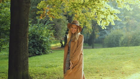 Cheerful-blonde-young-woman-wearing-hat-and-coat-running-and-whirling-in-the-park-in-autumn