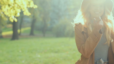 Close-up-view-of-blonde-young-woman-holding-her-hat-and-smiling-at-the-camera-in-the-park-un-autumn