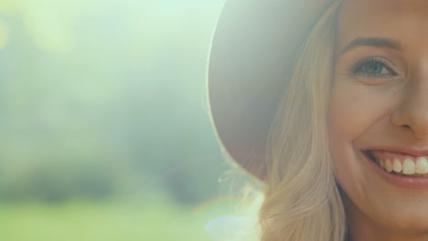 Close-up-view-of-half-face-of-blonde-young-woman-wearing-a-hat--and-smiling-cheerfully-to-the-camera-in-the-park-in-autumn