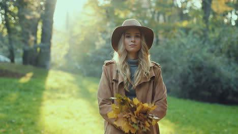 Cheerful-stylish-Caucasian-young-woman-wearing-a-hat-and-coat-having-fun-in-the-park,-throwing-autumn-yellow-leaves-and-looking-at-camera