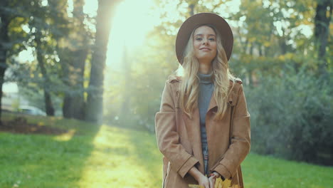 Cheerful-stylish-Caucasian-young-woman-wearing-a-hat-and-coat-having-fun-in-the-park,-throwing-autumn-yellow-leaves-and-looking-at-camera