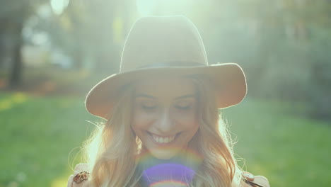 Caucasian-young-blonde-woman-wearing-a-hat-smiling-and-looking-at-camera-while-holding-autumn-yellow-leaves-in-the-park