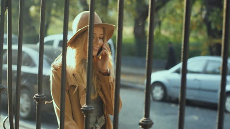 Cheerful-smiled-blonde-young-woman-weating-a-hat-and-coat-walking-behind-a-fence-and-talking-on-the-phone-in-the-street