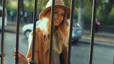 Cheerful-smiled-blonde-young-woman-weating-a-hat-and-coat-walking-behind-a-fence-in-the-street