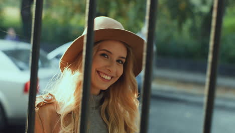 Cheerful-smiled-blonde-young-woman-weating-a-hat-and-coat-walking-behind-a-fence-while-looking-at-camera-in-the-street