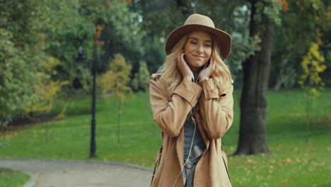 Caucasian-young-blonde-woman-putting-the-headphones-on-and-listening-to-the-music-while-walking-in-the-park-in-autumn