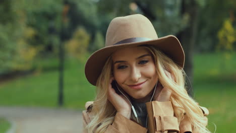 Close-up-view-of-caucasian-young-blonde-woman-putting-the-headphones-on-and-listening-to-the-music-while-walking-in-the-park-in-autumn