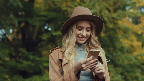 Kaukasische-Junge-Blonde-Frau,-Die-Einen-Hut-Trägt-Und-Im-Herbst-Eine-Sms-Auf-Dem-Smartphone-Im-Park-Schreibt