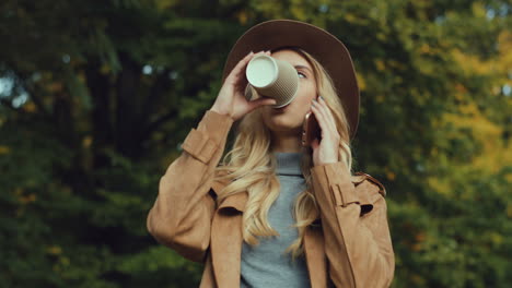 Nahaufnahme-Einer-Kaukasischen-Jungen-Blonden-Frau,-Die-Einen-Hut-Trägt-Und-Telefoniert-Und-Im-Herbst-Kaffee-Trinkt,-Um-In-Den-Park-Zu-Gehen