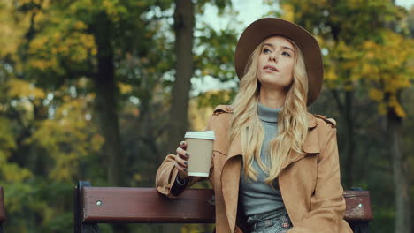 Caucasian-blonde-caucasian-woman-sitting-on-the-bench-in-a-park,-drinking-coffee-while-waiting-for-somebody-and-looking-at-the-watch