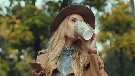 Kaukasische-Junge-Blonde-Frau-Mit-Hut-Und-Kopfhörern,-Die-Musik-Auf-Dem-Smartphone-Hört-Und-Kaffee-In-Einem-Schönen-Herbstpark-Trinkt
