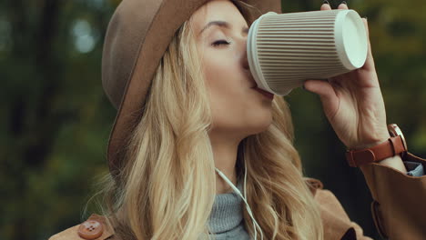 Close-up-view-of-caucasian-young-blonde-woman-in-a-hat-and-headphones-listening-the-music-on-the-smartphone-and-drinking-coffee-in-a-beautiful-autumn-park