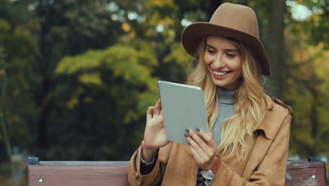Caucasian-blonde-caucasian-woman-sitting-on-the-bench-and-using-a-tablet-in-the-park-in-autumn